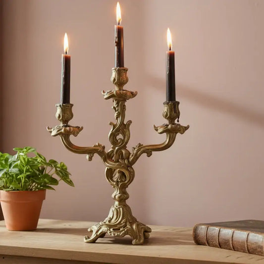Decorative brass candelabrum with three lit candles on a wooden surface, next to a potted plant and an old book.