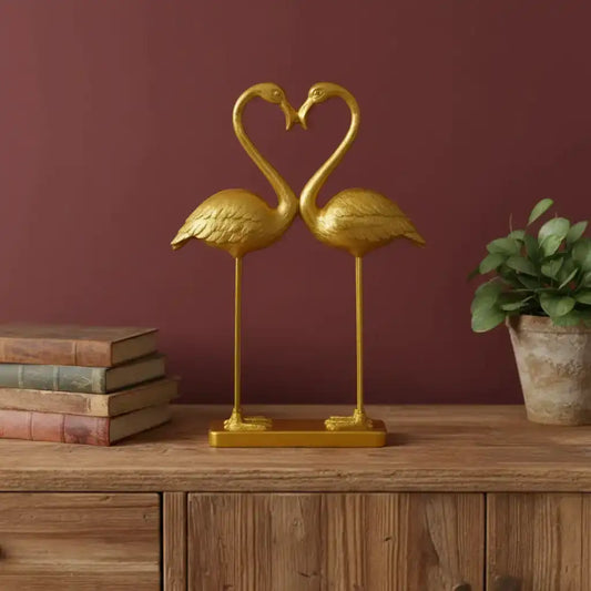 Gold flamingo sculpture on a wooden surface with a plant and books in the background.