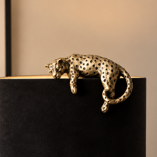 A metallic leopard figurine with a perforated, textured finish in a gold/bronze tone, resting across the top of a black cylindrical lamp base against a warm beige background.
