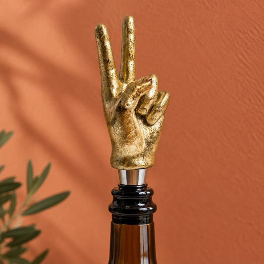 A gold hand-shaped bottle stopper on a dark brown glass bottle, set against a warm terracotta background with soft leaf shadows.