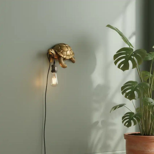 Gold turtle-shaped wall lamp with a lit bulb on a light gray wall, next to a potted plant.