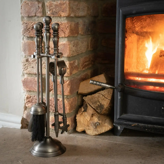 Set of metal fireplace tools next to a lit wood stove with logs in front of a brick wall.