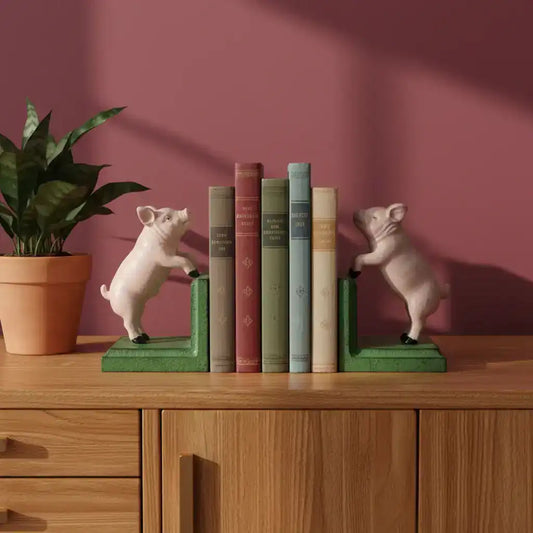 Pig-shaped bookends on a wooden shelf with books and a plant against a pink wall.