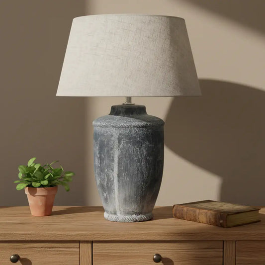Decorative lamp with textured base and beige shade in a room setting with framed artwork and mugs.