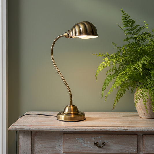 A brass gooseneck table lamp with a ribbed dome shade sits on a distressed light wood console table next to a potted green fern against a sage green wall.