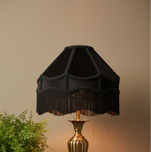 A black fabric lampshade with decorative trim and fringe, mounted on a gold-toned lamp base against a warm beige wall, with a small green plant at the lower left.