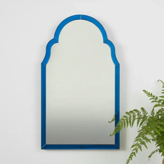 Arched mirror with a blue frame on a white wall with a plant to the right.