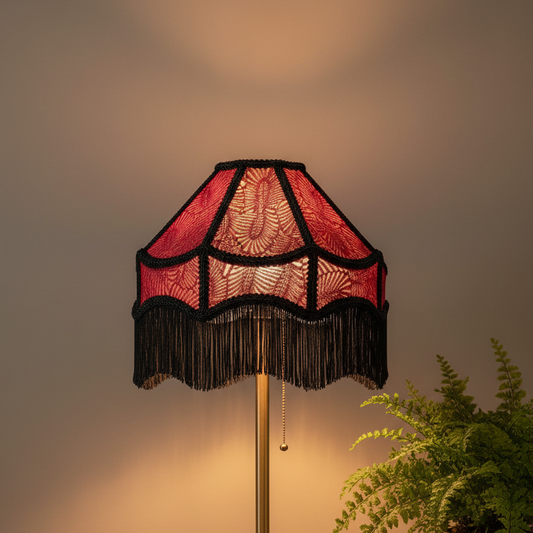 A vintage-style floor lamp with a red and black patterned fringed shade, illuminated to cast a warm glow against a neutral background with a green fern in the foreground.