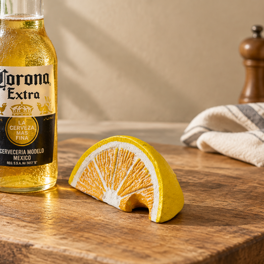 A ceramic orange slice candle holder sits on a wooden table next to a Corona Extra bottle, with a neutral background and a folded cloth.