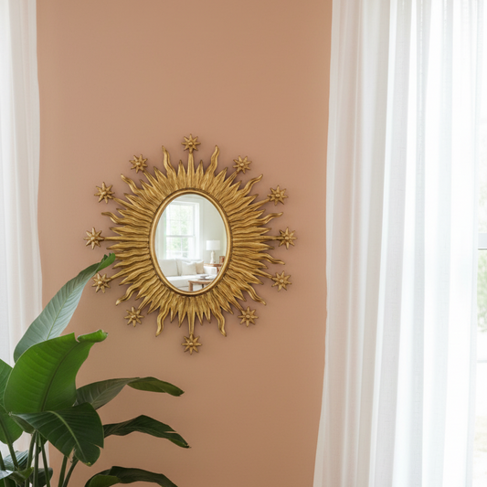 Decorative gold sunburst mirror on a beige wall with a plant and window in the foreground.