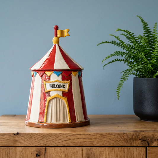 A ceramic circus tent-shaped container with a red and white striped pattern, a yellow 'WELCOME' sign, and a small yellow flag on top, displayed on a wooden surface next to a potted fern against a blue wall.