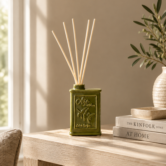 An olive green reed diffuser bottle with embossed olive branch details and natural reed sticks, styled on a light wood table next to stacked books and a vase in a softly lit room.