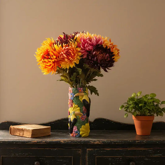 Colorful vase with flowers on a wooden surface next to a book and small potted plant.