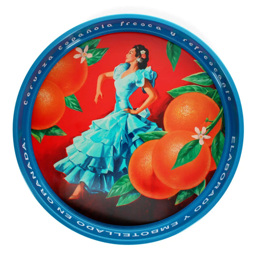 A round metal tin lid featuring a vibrant illustration of a woman in a light blue dress dancing among large oranges and green leaves on a red background, with a blue rim inscribed with Spanish text.
