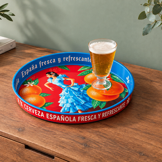 A round decorative serving tray with a red and blue Spanish-themed design, featuring an illustration of a woman in a blue dress, oranges, and the text 'Cerveza Española Fresca y Refrescante'. A glass of beer is placed on top of the tray, which sits on a wooden table against a muted green wall.