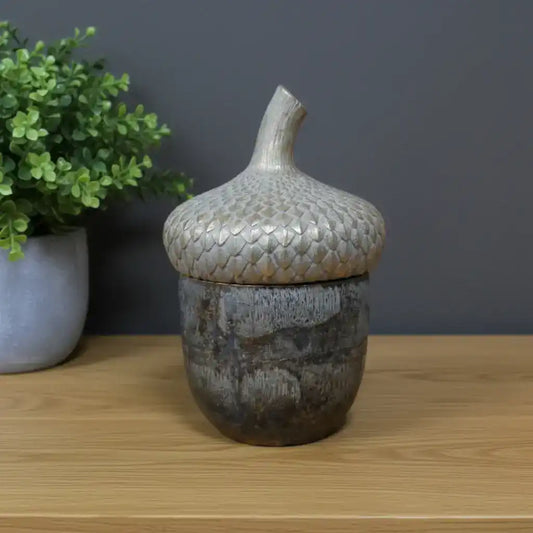 Decorative ceramic acorn-shaped container on a wooden surface with a plant and framed picture in the background.