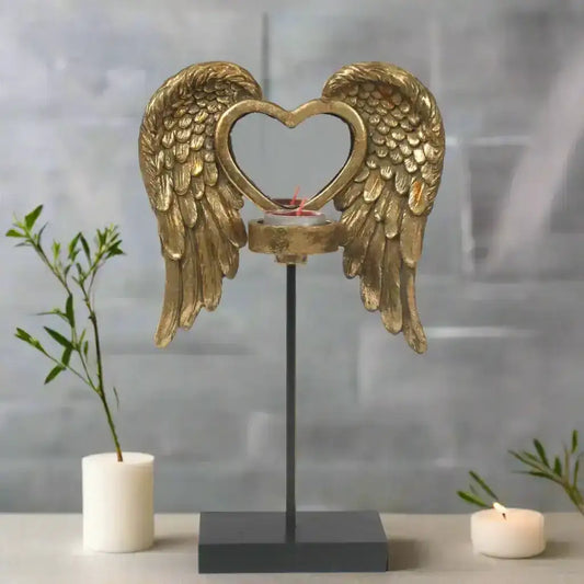 Decorative item with gold angel wings and a heart-shaped design on a stand, with candles and plants in the background.