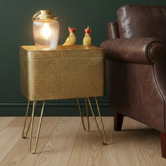 Gold textured side table with a lamp and decorative items next to a brown leather armchair.