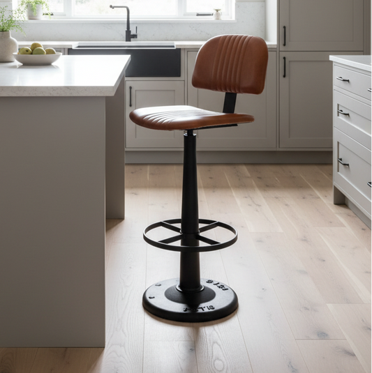 A modern kitchen bar stool with a brown leather seat and backrest, set in a bright kitchen with light wood floors and gray cabinets.