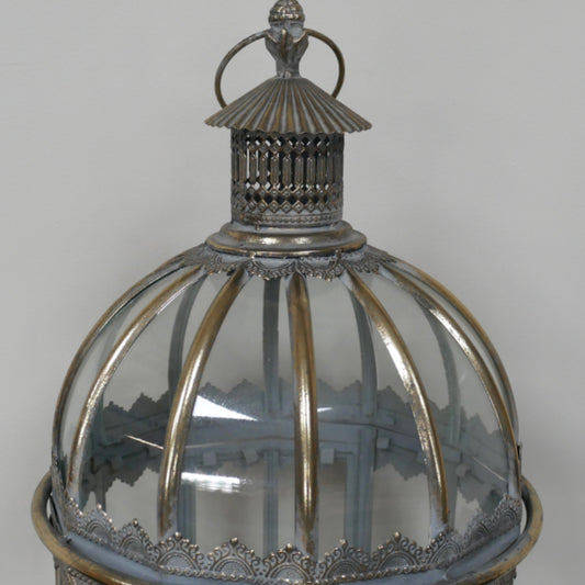 A close-up of an ornate vintage lantern with a clear ribbed glass dome, silver-toned metal frame, and decorative top finial against a neutral gray background.