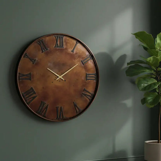 Bronze wall clock with Roman numerals on a dark green wall with a plant to the right.