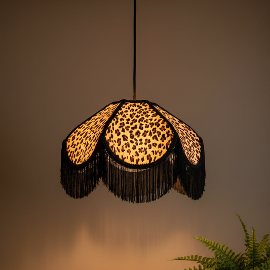 A hanging pendant lamp with a leopard-print fabric shade and long black fringe, shown lit against a warm neutral wall with a small plant visible in the lower right corner.