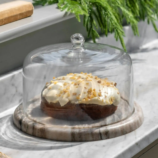 Decorative cake with white frosting and gold sprinkles under a glass cloche on a marble surface.