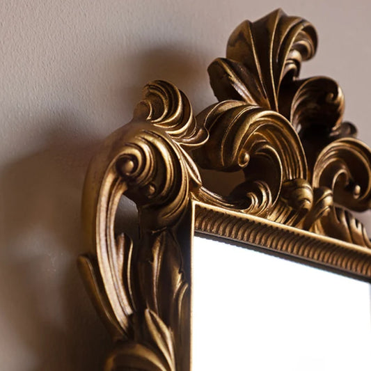 Close-up of an ornate gold mirror frame with carved baroque-style scrollwork and a reflective white mirror surface, mounted against a beige wall.