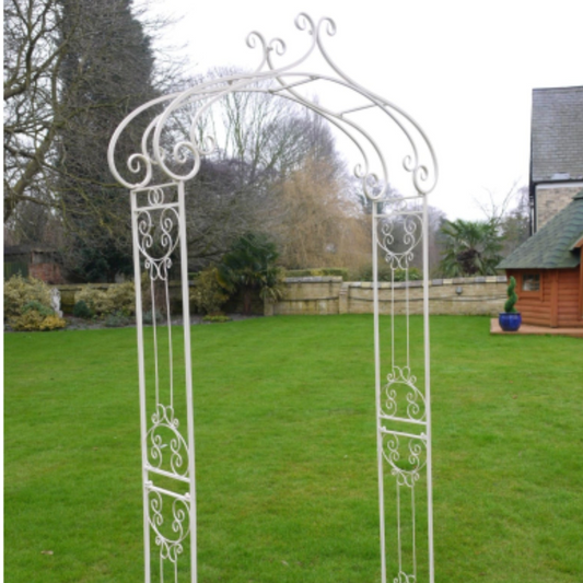 Decorative white metal archway in a garden setting with trees and a building in the background.