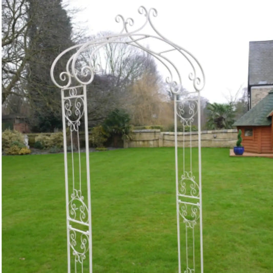 Decorative white metal archway in a garden setting with trees and a building in the background.