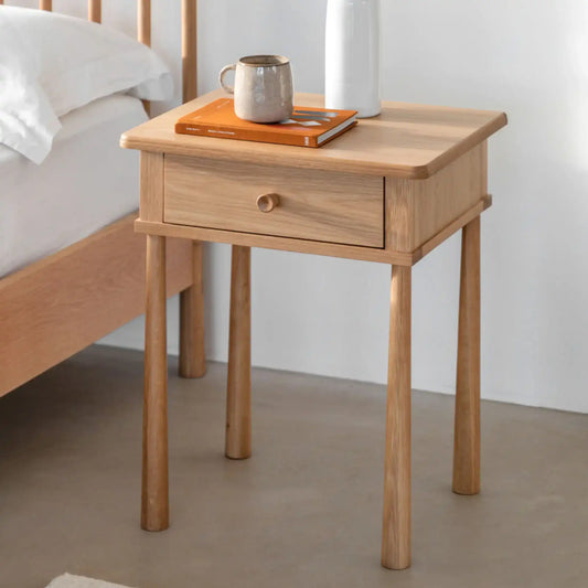 Wooden side table with a drawer next to a bed, featuring a mug and book on top.