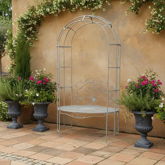 Metal garden arch with bench against a beige wall with potted plants.