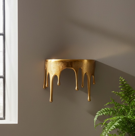 Gold wall-mounted lamp with a modern design on a gray wall next to a window and plant.