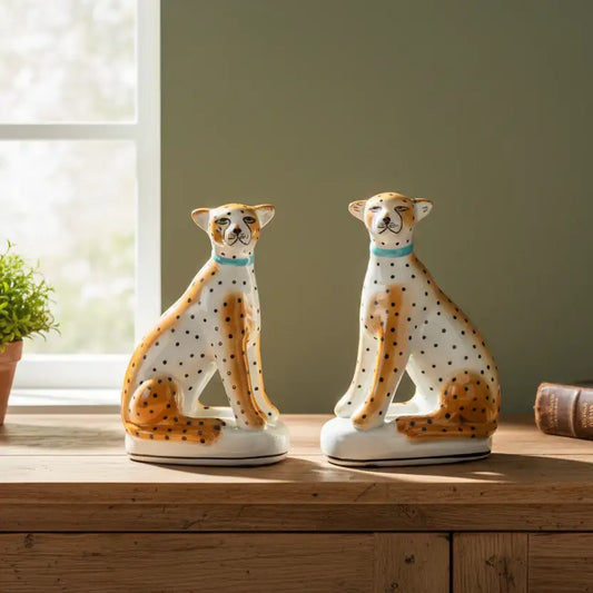 Two ceramic cheetah figurines on a wooden surface with a window and plant in the background.
