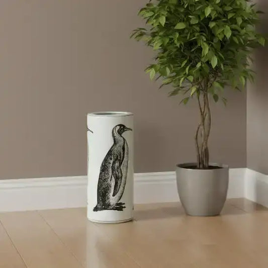 Cylindrical vase with penguin design next to a potted plant on a wooden floor.