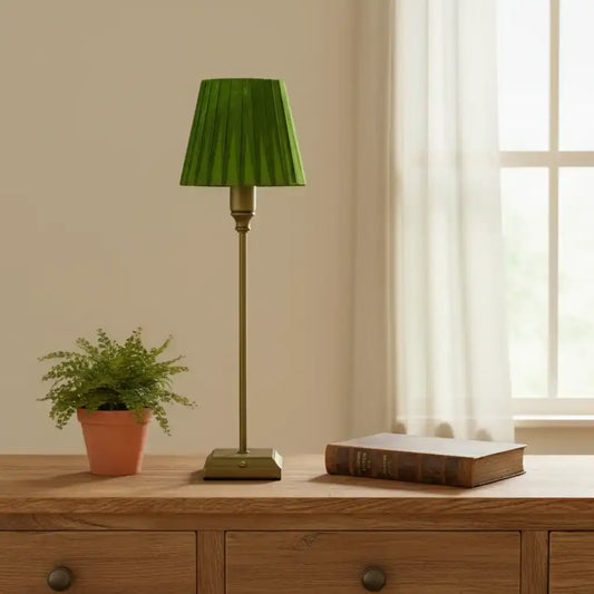 Green lamp on a wooden table with a plant and book, in a room with a window and beige wall.