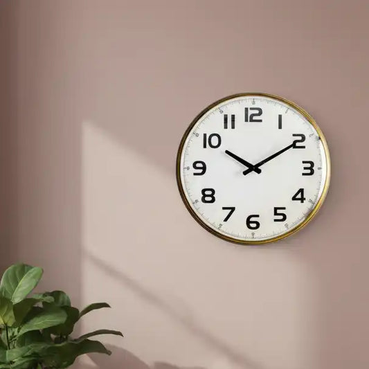 Round wall clock with gold frame on a beige wall, next to a plant.