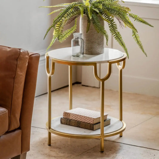 Small round marble table with gold frame next to a brown leather chair, with a plant in the background.