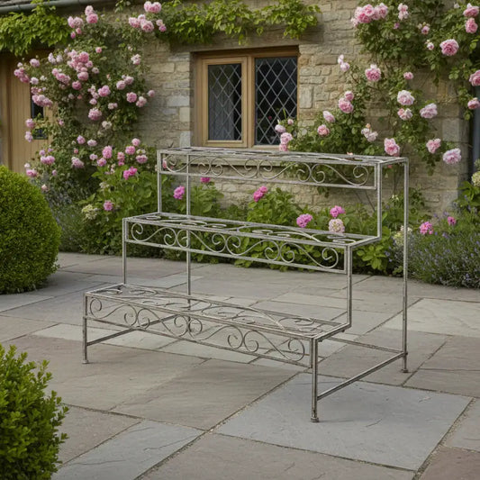 Wrought iron plant stand in a garden setting with pink flowers and a stone building.