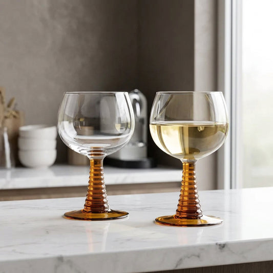 Two clear wine glasses with ribbed amber-brown stems and round bases, placed on a beige-and-white checkered tablecloth. One glass is empty, and the other contains white wine with a dark olive on top.