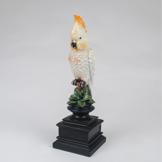 A decorative figurine of a white cockatoo with a bright orange-yellow crest perched on a black pedestal with green leaves, photographed against a neutral light gray background.