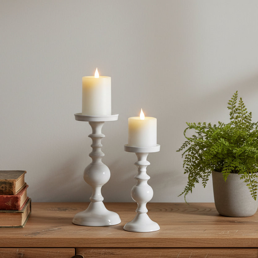 Two white candlesticks with lit candles on a wooden surface, accompanied by a potted plant and books.