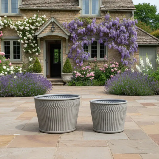Two gray outdoor planters on a stone patio with a garden and house in the background.
