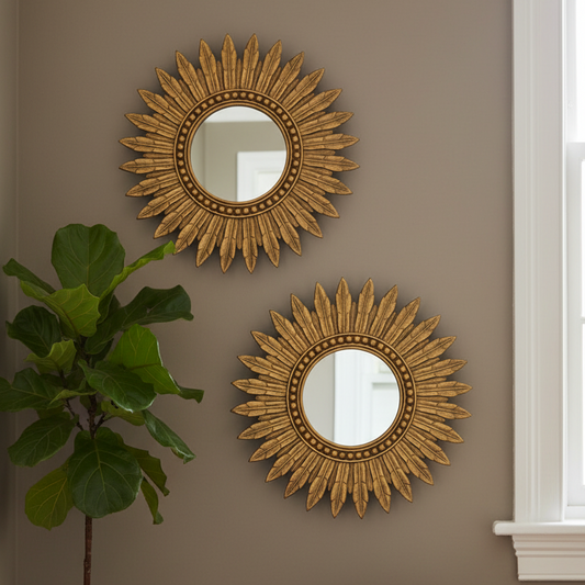 Two decorative gold sunburst mirrors mounted on a taupe wall, with a green fiddle-leaf fig plant on the left and a white window frame on the right.