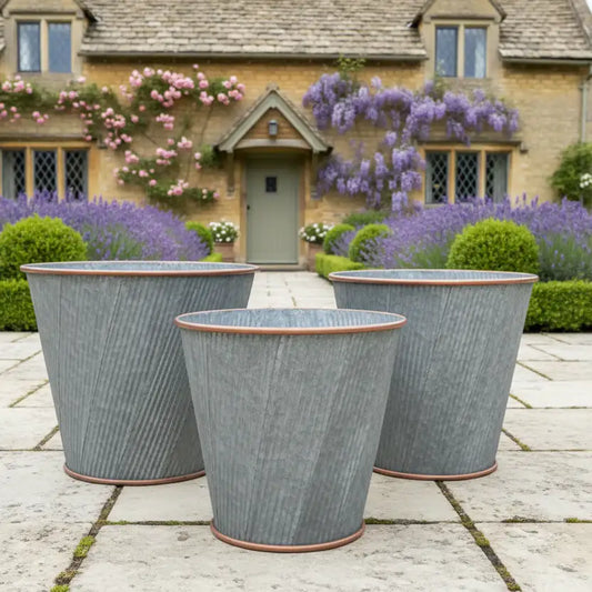 Three gray metal planters on a stone patio with a house and flowers in the background