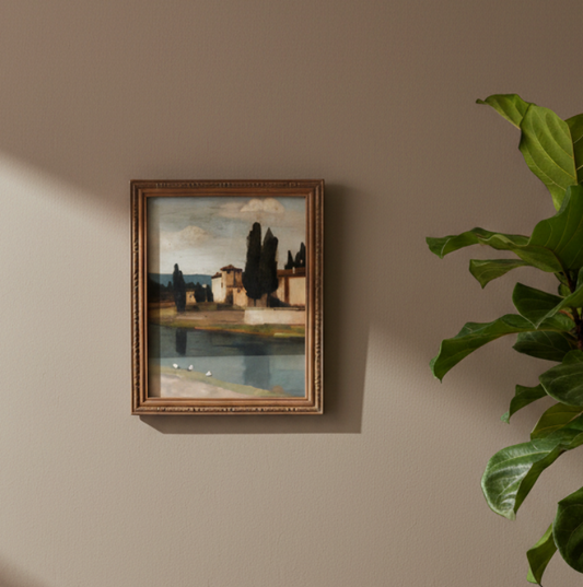 A framed landscape painting hanging on a beige wall, styled next to a green houseplant with sunlight casting shadows across the wall.
