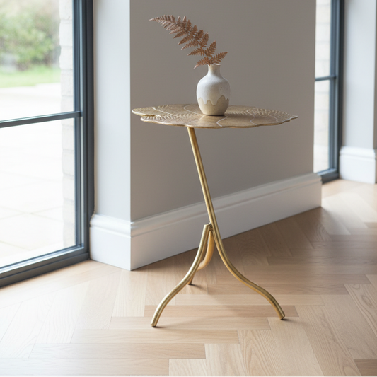 A gold metal leaf-shaped side table with a small white vase containing a dried fern, set on a light wood herringbone floor in a bright room with large windows.