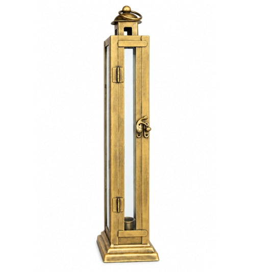 A tall, narrow brass lantern-style candle holder with a lit red pillar candle inside, displayed on a distressed white wooden cabinet against a neutral gray wall.