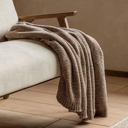 Brown knitted blanket draped over a beige armchair on a wooden floor.