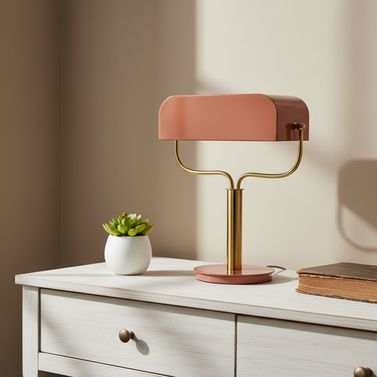 Modern table lamp with a pink shade and gold base on a white surface with a plant and book.